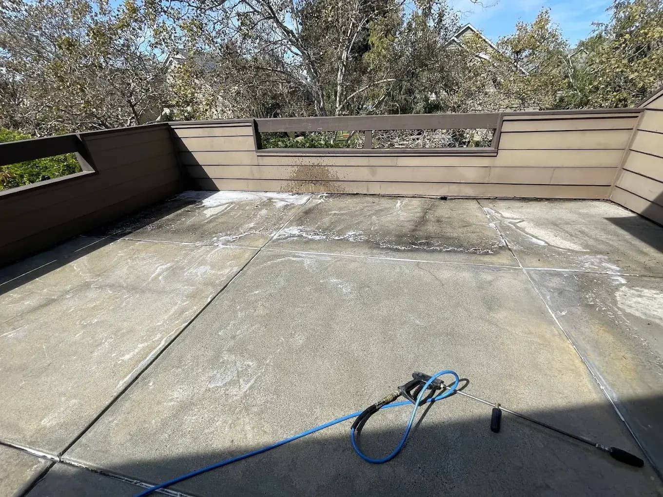 Large outdoor concrete patio being professionally power washed showing a clean 8x8 foot section, water runoff, lifted dirt, and trees in the background.