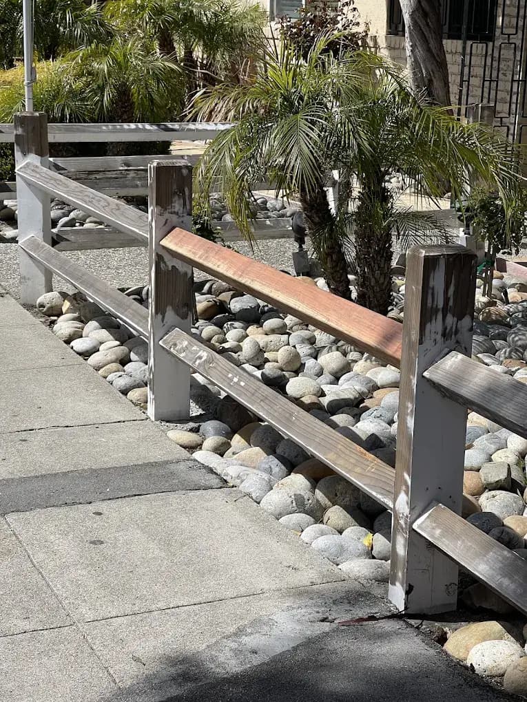 Before and after power washing of a wooden railing by a sidewalk, showing the top rail freshly cleaned to natural wood while the bottom rail remains dark and dirty.